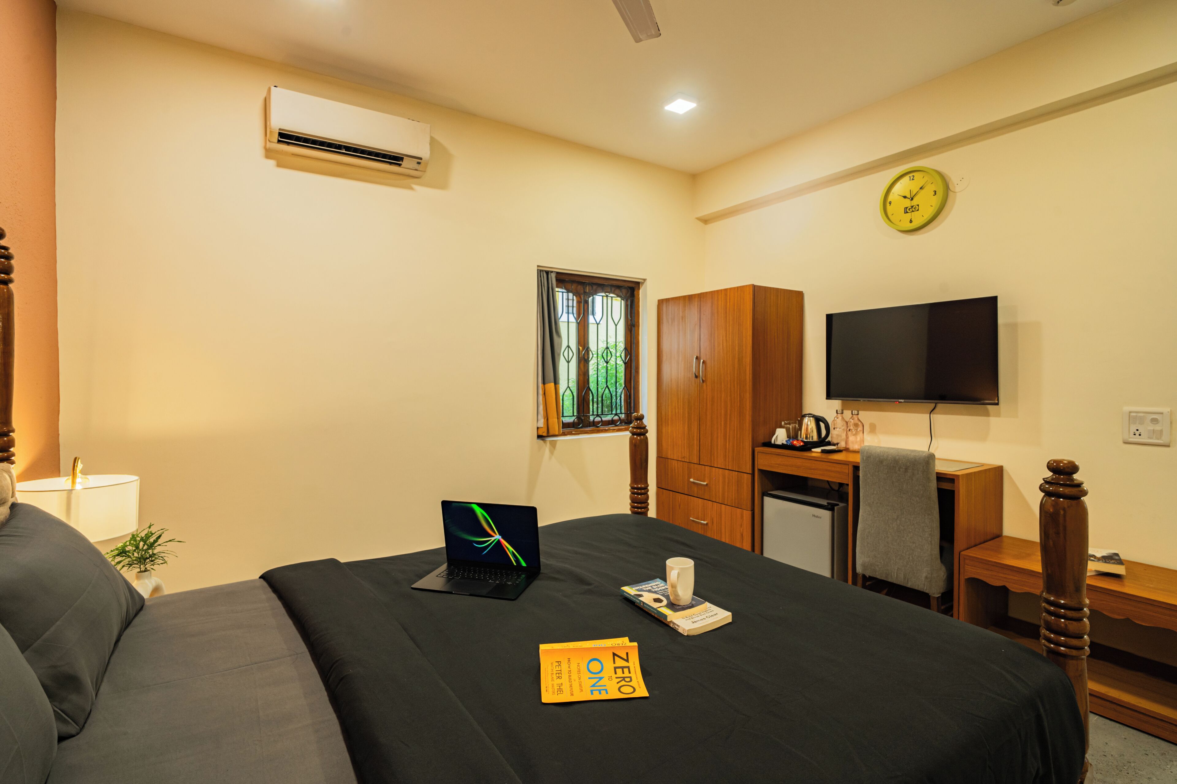 A hotel room with a bed, a desk, a television, a clock, and an air conditioning unit.