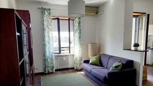 A living room with a sofa, a radiator, a window with curtains, a lamp, and a bookshelf.
