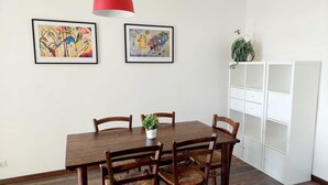A dining area with a wooden table and chairs, two framed abstract paintings, a red pendant light, and a white shelving unit.