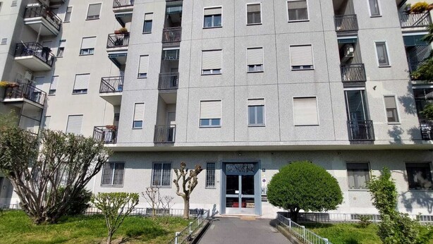 A multi-story residential building with balconies, a central entrance, and a small tree in the foreground.