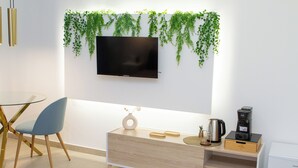En moderne stue med et fladskærms-tv monteret på væggen, et spisebord med glasplade, en blå stol, et træsofabord med kaffemaskine, og en lille plante hængende over.