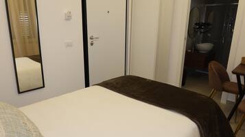 Habitació d'hotel amb llit, mirall, porta i bany visible per una porta oberta.