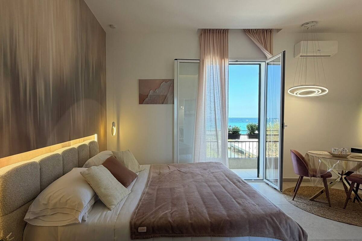 A modern bedroom with a large bed, a view of the sea, and a dining area with a table and chairs.