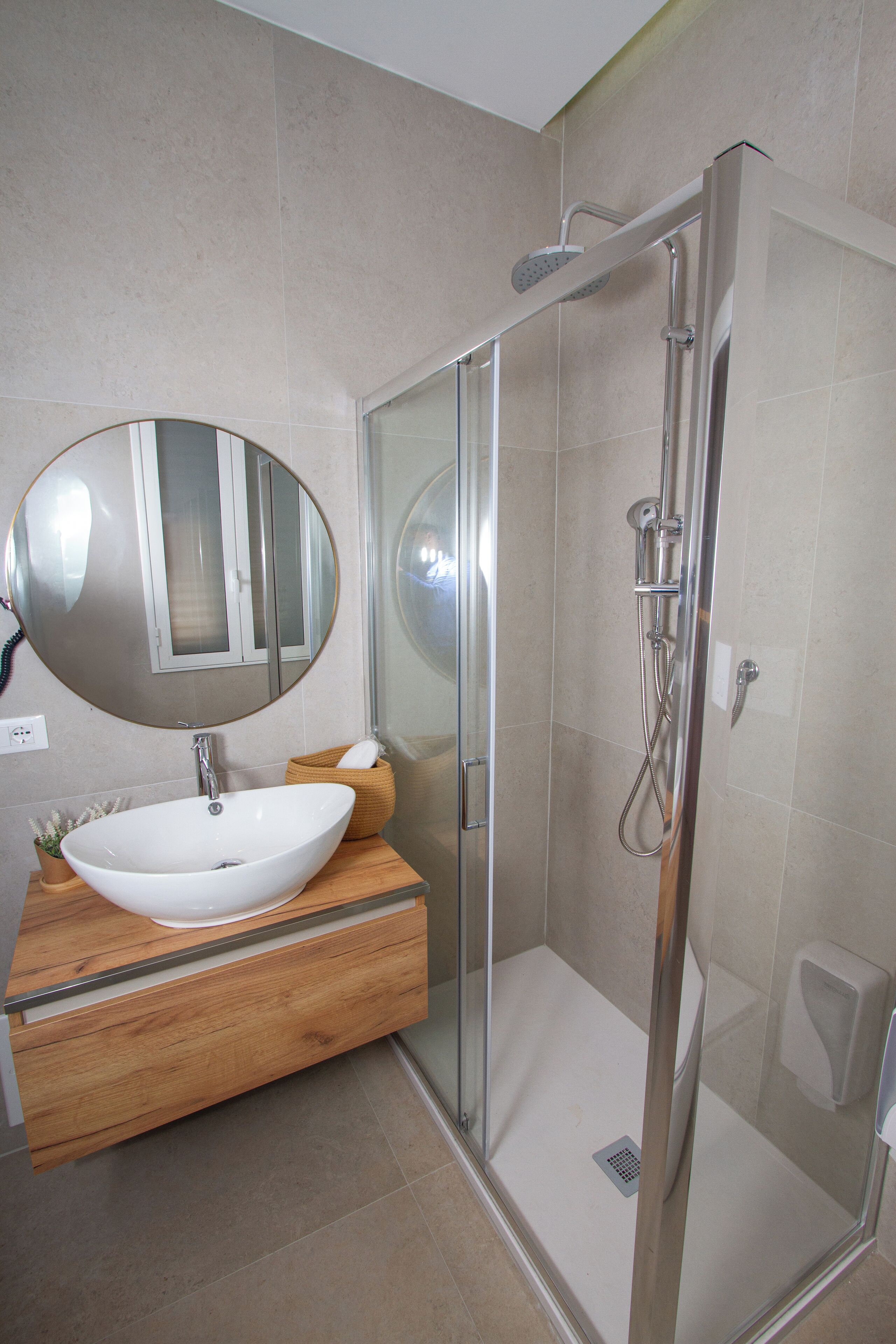 Un bagno moderno con cabina doccia in vetro, uno specchio rotondo e un mobile bagno in legno con lavandino bianco.