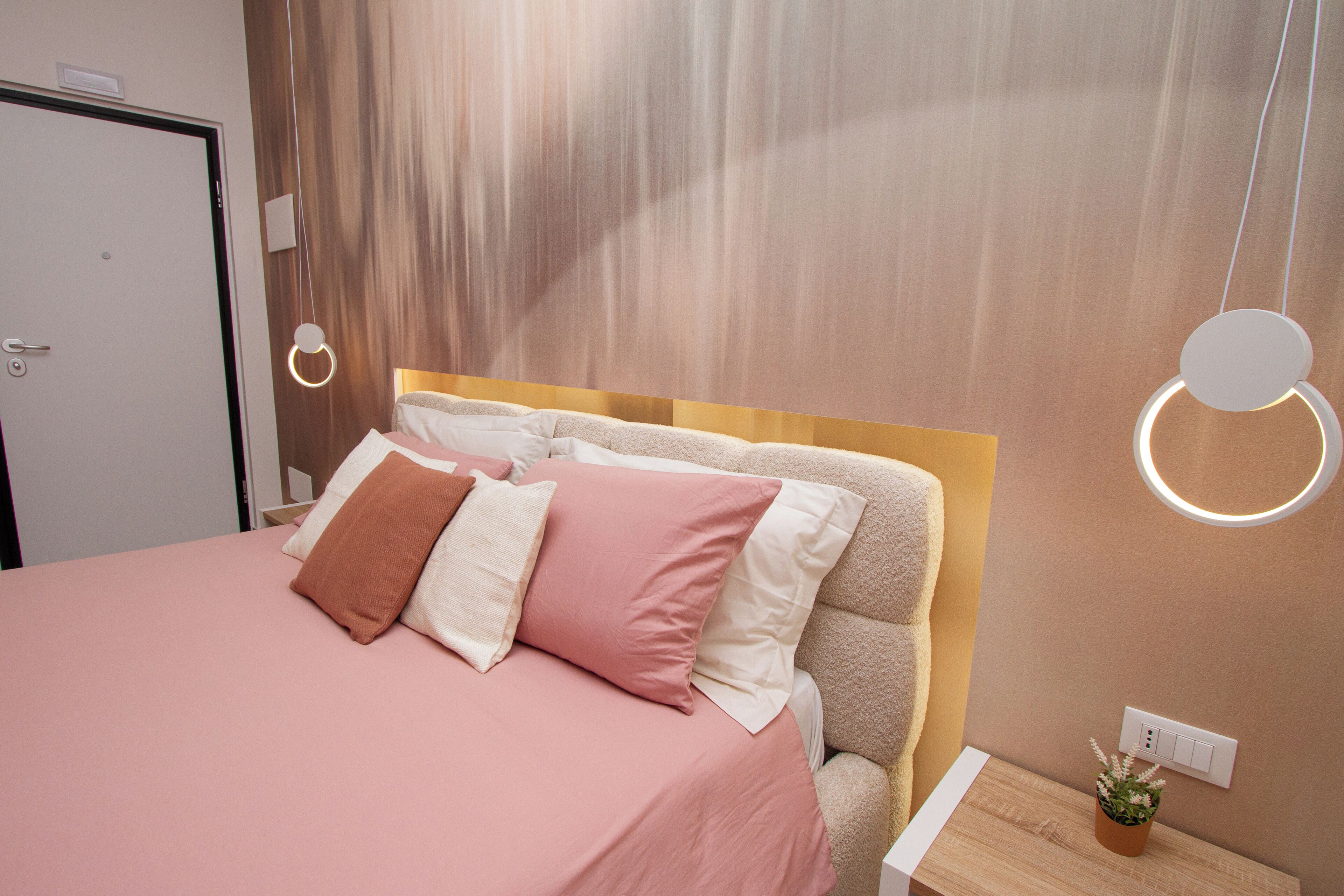 A neatly made bed with a pink bedspread and multiple pillows, a wooden headboard, and a bedside table with a small plant.