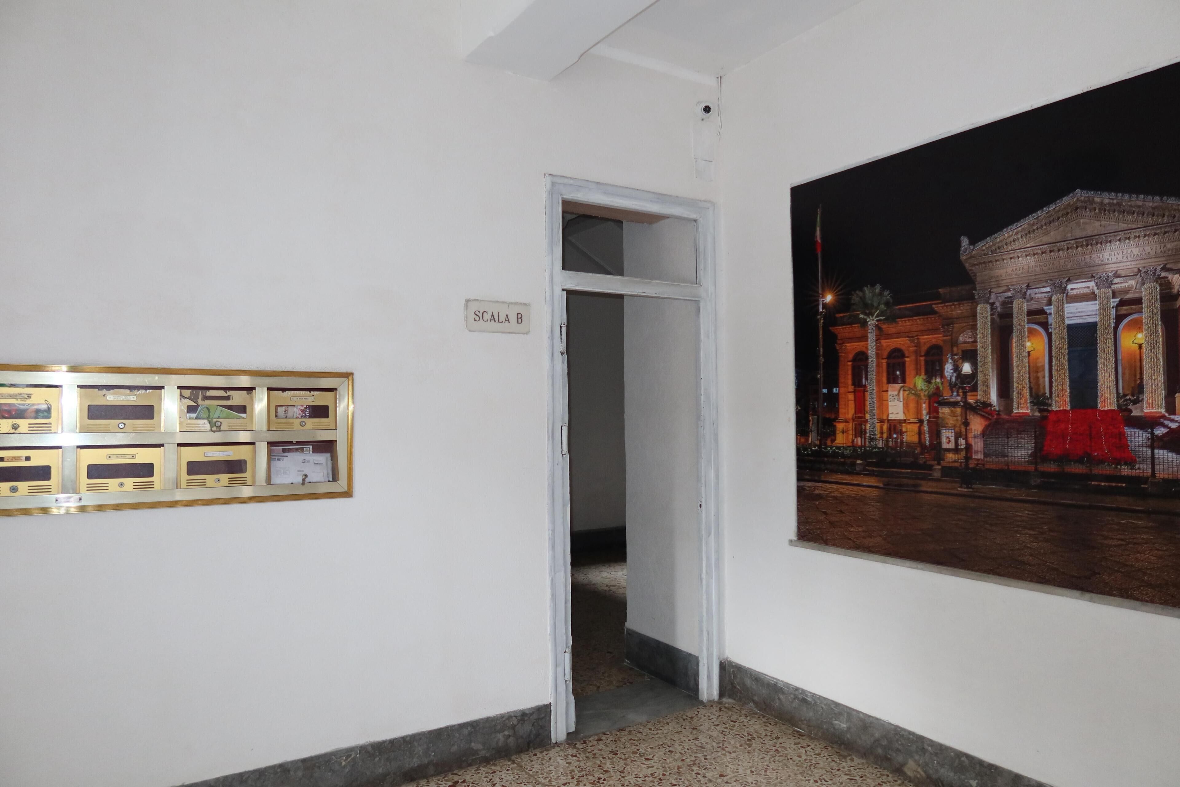 Un corridoio con una porta recante la scritta "SCALA B" che dà accesso a una stanza con un grande quadro raffigurante un edificio di notte.