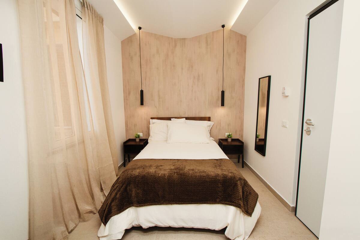 A modern bedroom with a large bed, two bedside tables, a nightstand, a mirror, and sheer curtains.