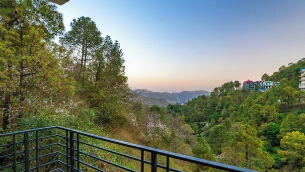 A balcony with a metal railing overlooking a forested hillside with buildings in the background. - The Hosteller Kasauli (Kasauli)