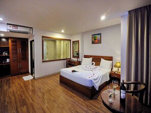 Habitación de hotel con piso de madera, cama con ropa blanca, mesita de noche con lámpara y una pequeña mesa redonda con una botella de vino y copas.