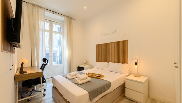 Habitación de hotel con cama, escritorio, silla, mesita de noche, televisor y ventana con cortinas.