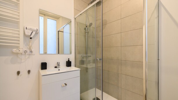 Une salle de bain moderne dotée d’une douche à parois de verre, d’un meuble-lavabo blanc avec un miroir et d’un sèche-cheveux fixé au mur.
