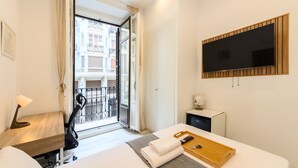 Une chambre d’hôtel comprenant un lit, un bureau, une télévision, un petit placard et un balcon avec vue sur les immeubles.