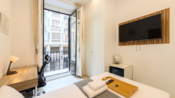 Une chambre d’hôtel comprenant un lit, un bureau, une télévision, un petit placard et un balcon avec vue sur les immeubles.