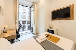 Ein Hotelzimmer mit Bett, Schreibtisch, Fernseher, kleiner Kommode und Balkon mit Blick auf Gebäude.