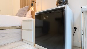 Ein kompakter Raum mit einem Bett, einem weißen Nachttisch, einem schwarzen Kühlschrank und Holzboden.