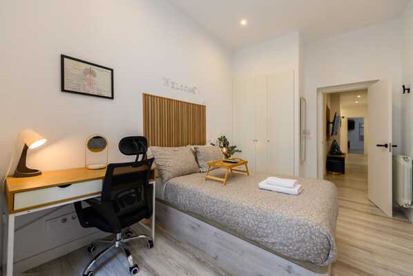 A modern bedroom with a wooden desk, a black office chair, a bed with a grey floral bedspread, a bedside table with a lamp, and a 'Welcome' sign on the wall.