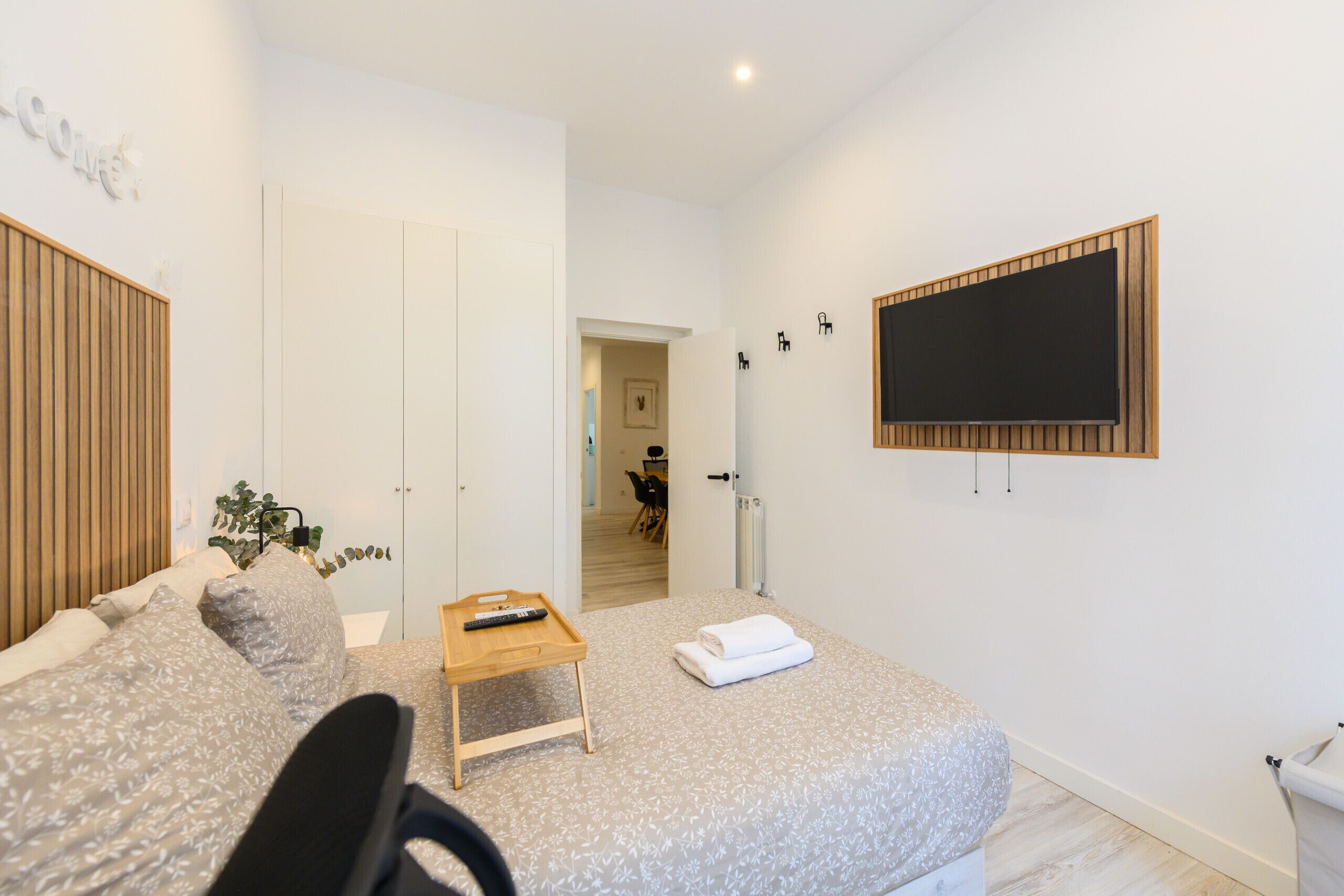 A modern bedroom with a bed, a small wooden table, a TV mounted on the wall, and a door leading to another room.
