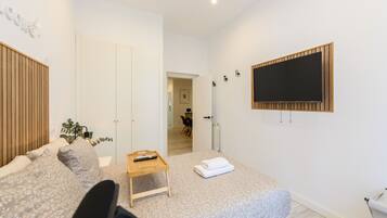 A modern bedroom with a bed, a small wooden table, a TV mounted on the wall, and a door leading to another room.