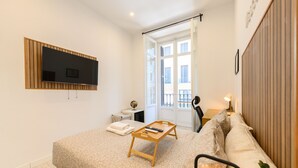 Une chambre moderne avec un lit, un téléviseur fixé au mur, une table de chevet avec une lampe et une fenêtre donnant sur des immeubles.