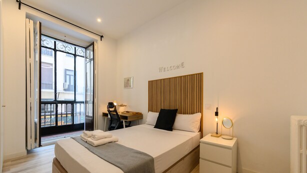A modern hotel room with a large bed, a desk with a chair, a bedside table with a lamp, and a view of the outside balcony. - Zurich: room with comfort and shared areas Gran Vía-Madrid (Madrid)