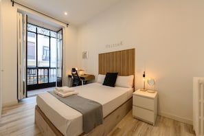 Ein modernes Hotelzimmer mit einem großen Bett, einem Schreibtisch mit Stuhl, einem Nachttisch mit Lampe und Blick auf den Balkon.