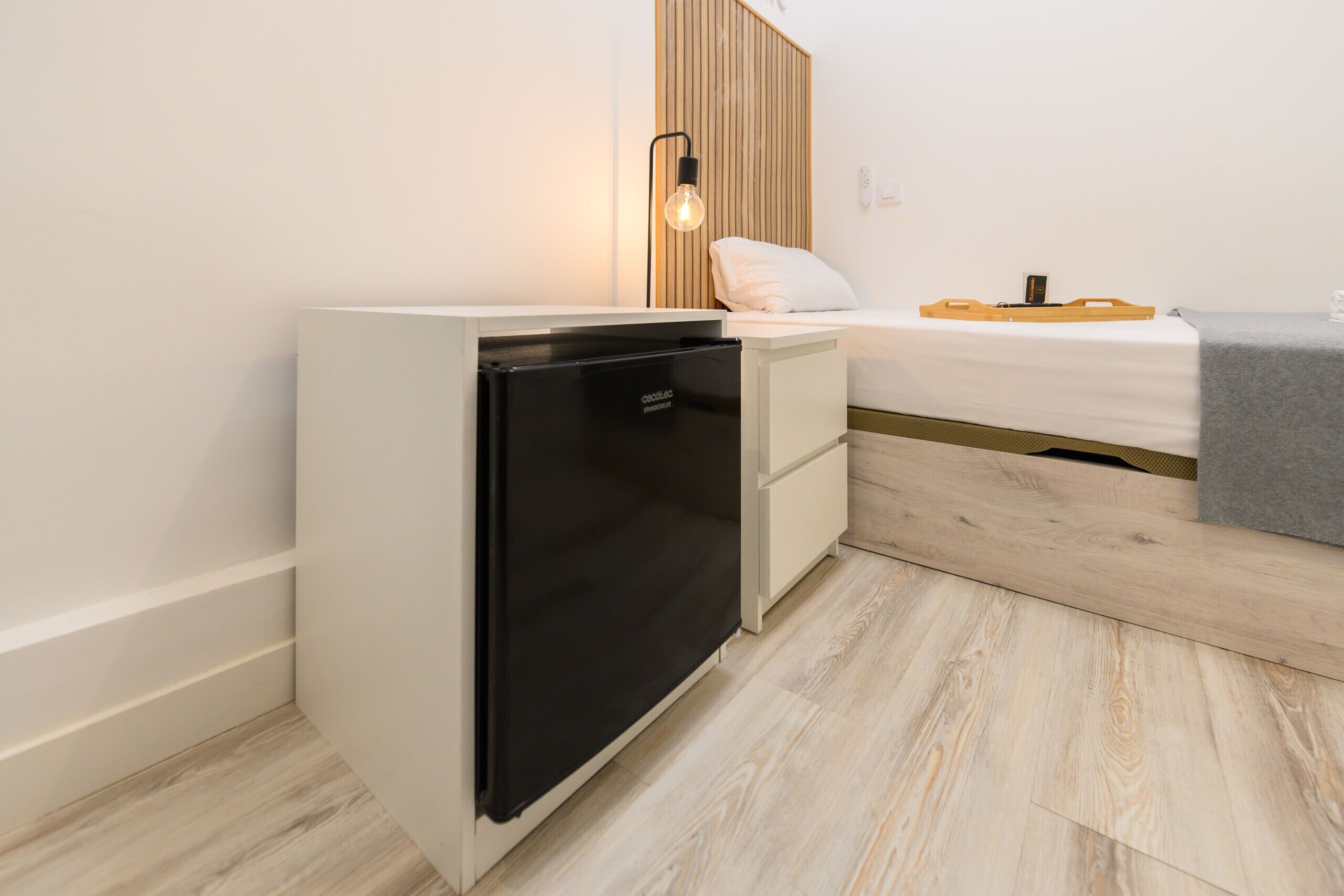 A compact room with a built-in black refrigerator, a small bedside table with a lamp, and a bed with a grey blanket.
