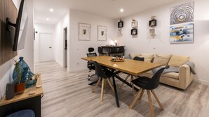 Un salon moderne avec une table à manger en bois, des chaises noires, un canapé beige et un téléviseur fixé au mur.