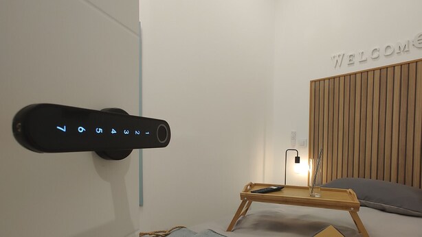Une chambre d’hôtel moderne équipée d’un lit, d’une table de chevet avec une télécommande, d’un panneau d’accueil et d’un panneau de commande fixé au mur.