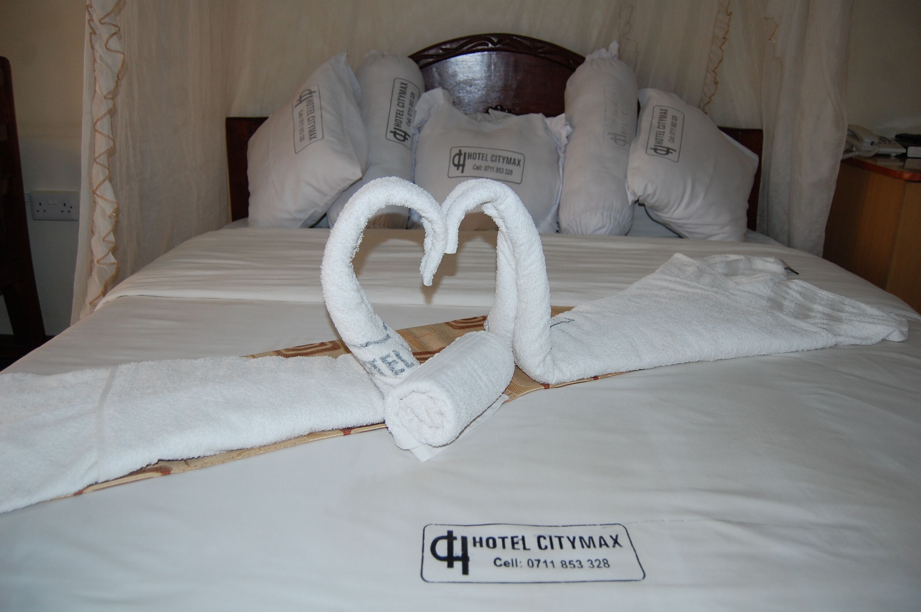 A hotel bed with a heart-shaped towel arrangement and pillows labeled "HOTEL CITYMAX."