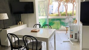 A modern living room with a white table, black chairs, a flat-screen TV, a standing fan, and a window with a view of buildings and trees.