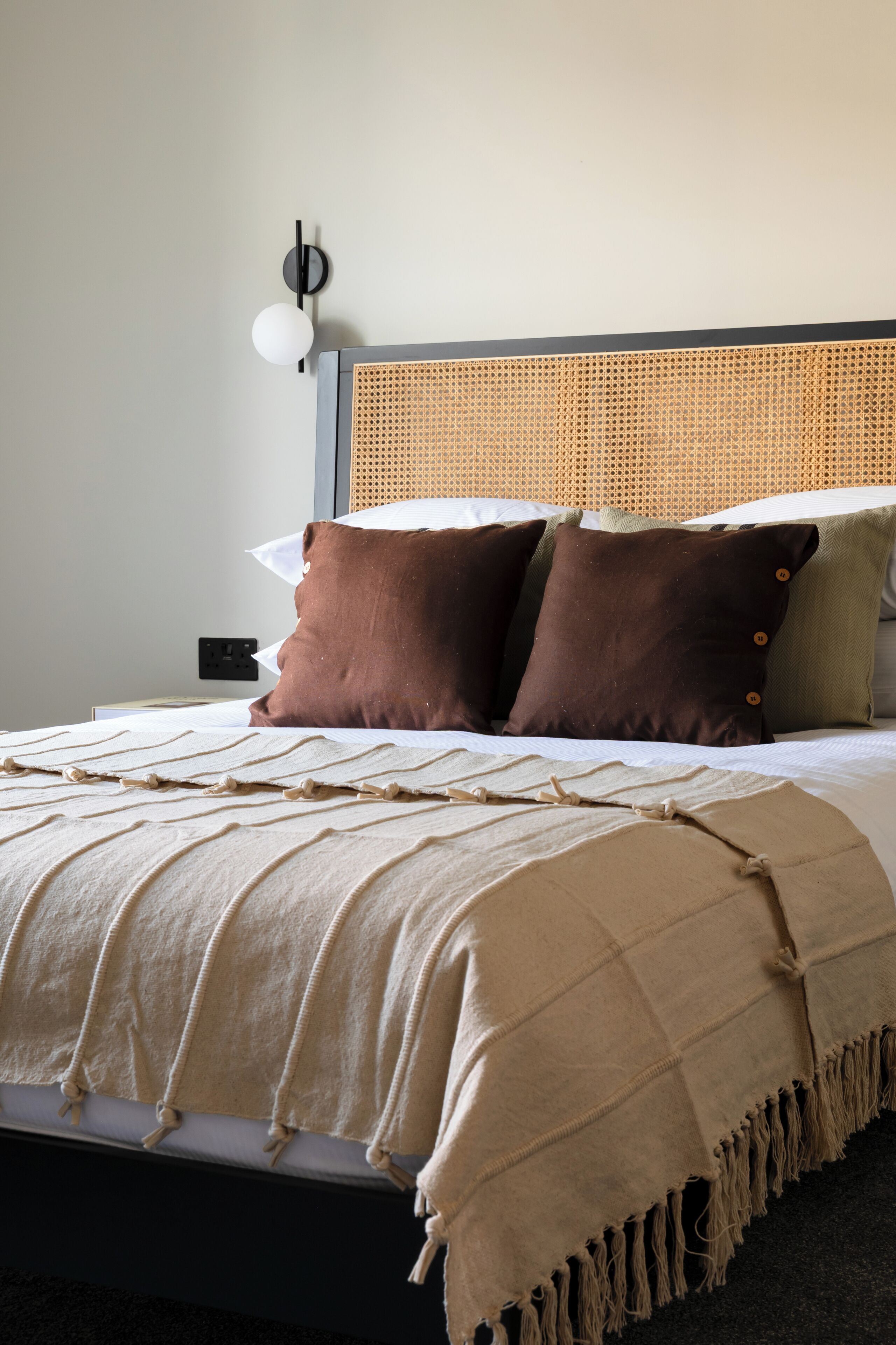 A neatly made bed with a beige blanket and pillows, a woven headboard, and a wall-mounted light fixture.