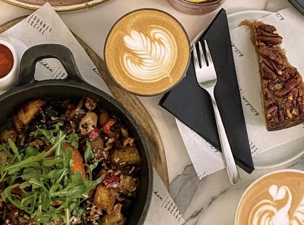 A meal with a dish of greens and potatoes, a cup of coffee with latte art, and a pecan-topped dessert on a plate.