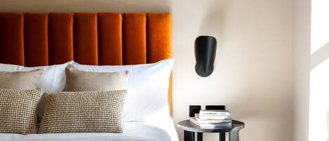 A neatly made bed with a tufted headboard, two patterned pillows, and a bedside table with a lamp and books.