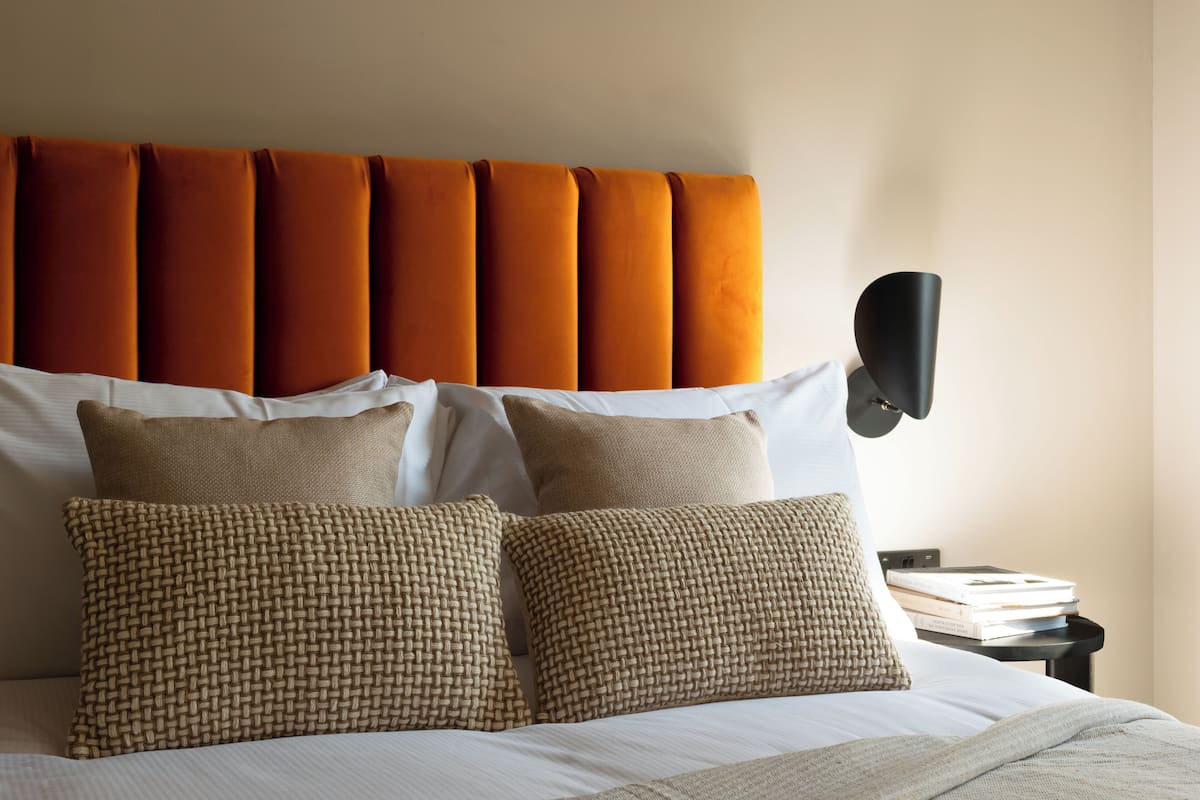 A neatly made bed with a headboard, several pillows, and a bedside table with a lamp and books.