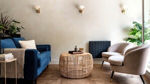 A modern living room with a blue sofa, a round wicker ottoman, a white armchair, and a black floor lamp. - Domain Boutique Rooms (Lancaster)
