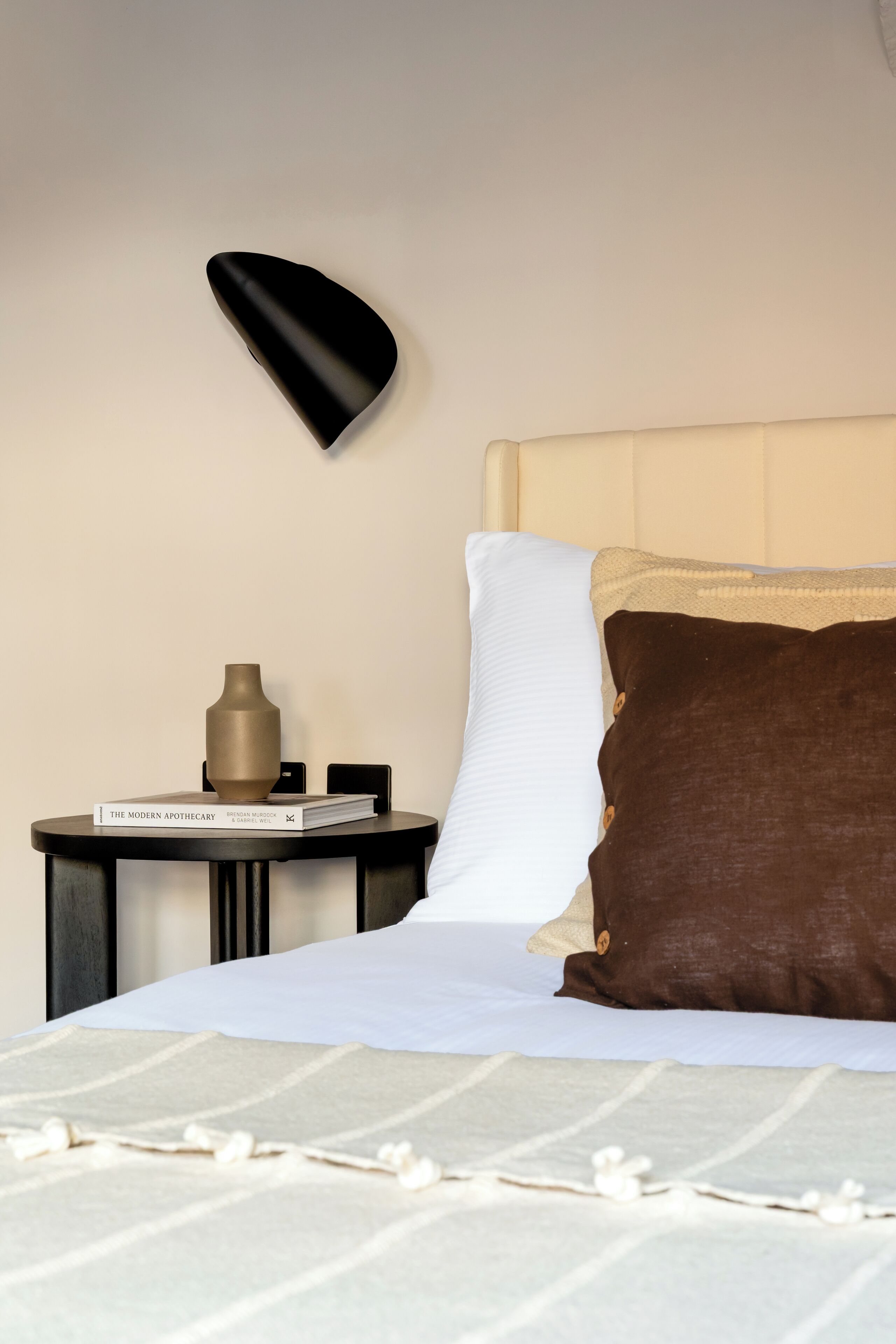 A neatly made bed with white and brown bedding, a bedside table with a book and a vase, and a wall-mounted lamp.