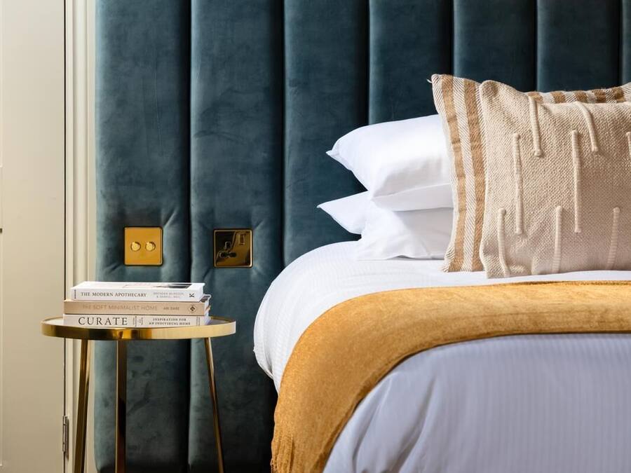 A neatly made bed with white linens and a mustard-colored throw blanket, a small bedside table with a stack of books, and a wall-mounted light fixture.