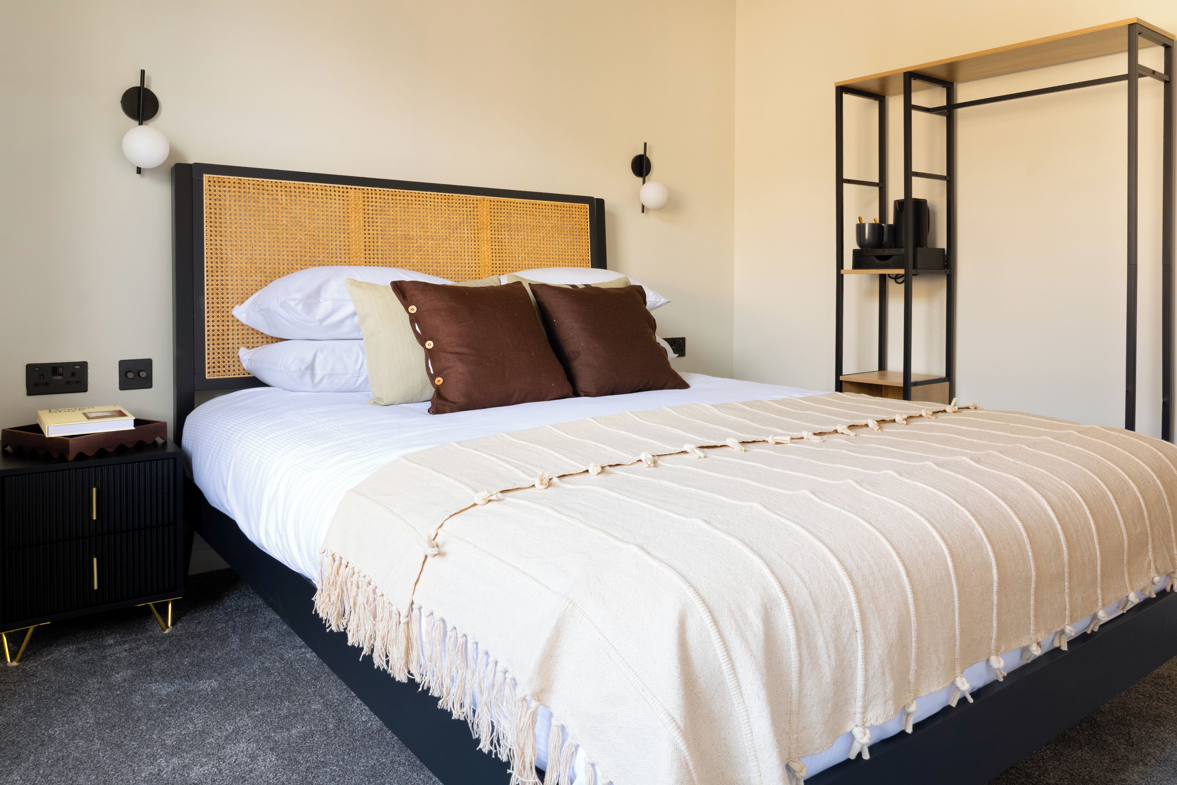 A neatly made bed with a striped blanket, a black nightstand with a phone, and a wall-mounted shelf with a lamp.