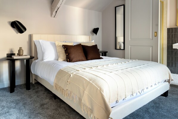 A neatly made bed with a white headboard, a beige throw blanket, and two brown pillows. There are two bedside tables with a book and a vase on one. The room has a gray carpet, a white wall, and a black and gold door. - Domain Boutique Rooms (Lancaster)