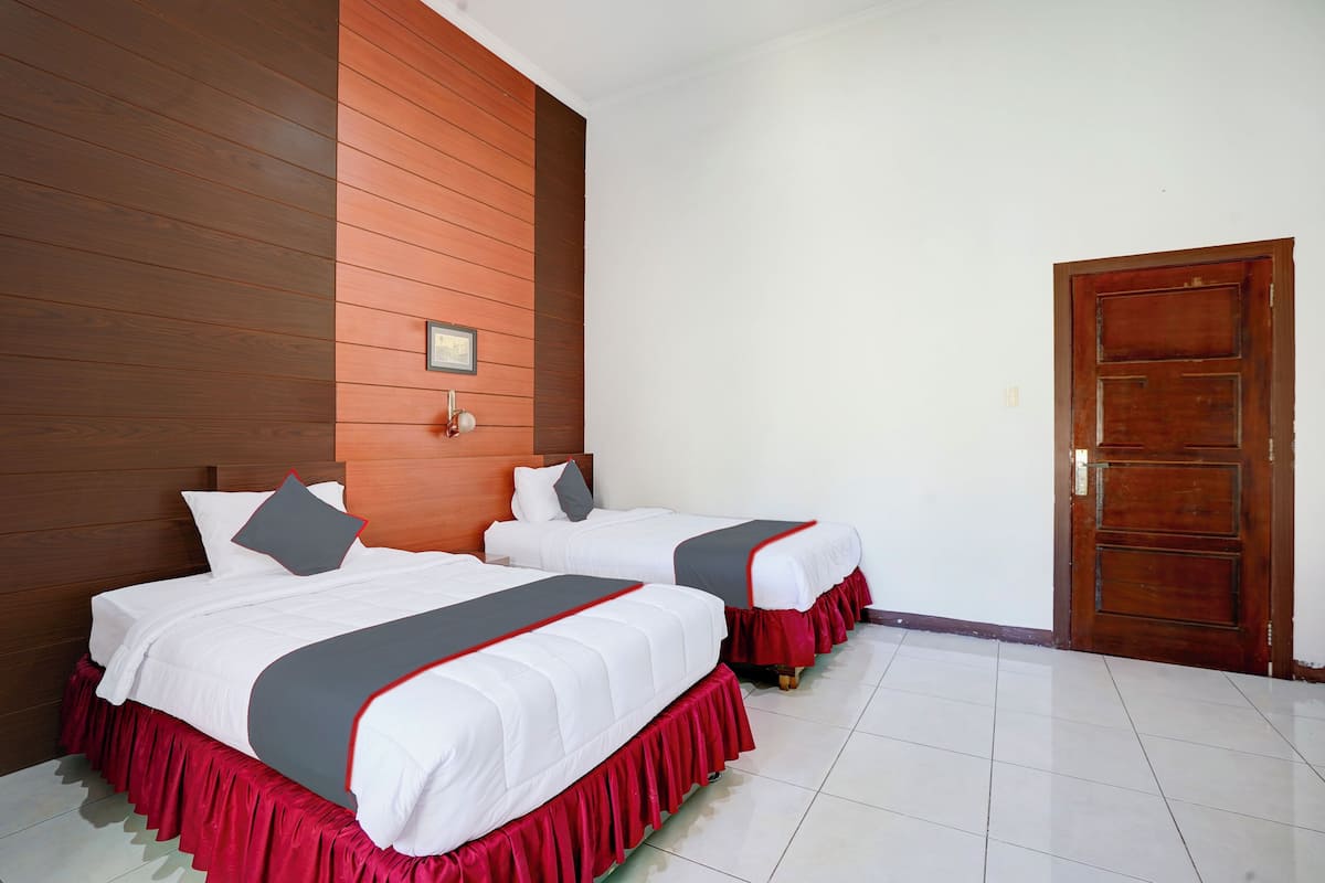 Une chambre d’hôtel avec deux lits, chacun agrémenté de draps blancs et de touches de couleur rouge, une tête de lit en bois, une porte en bois et un sol carrelé.