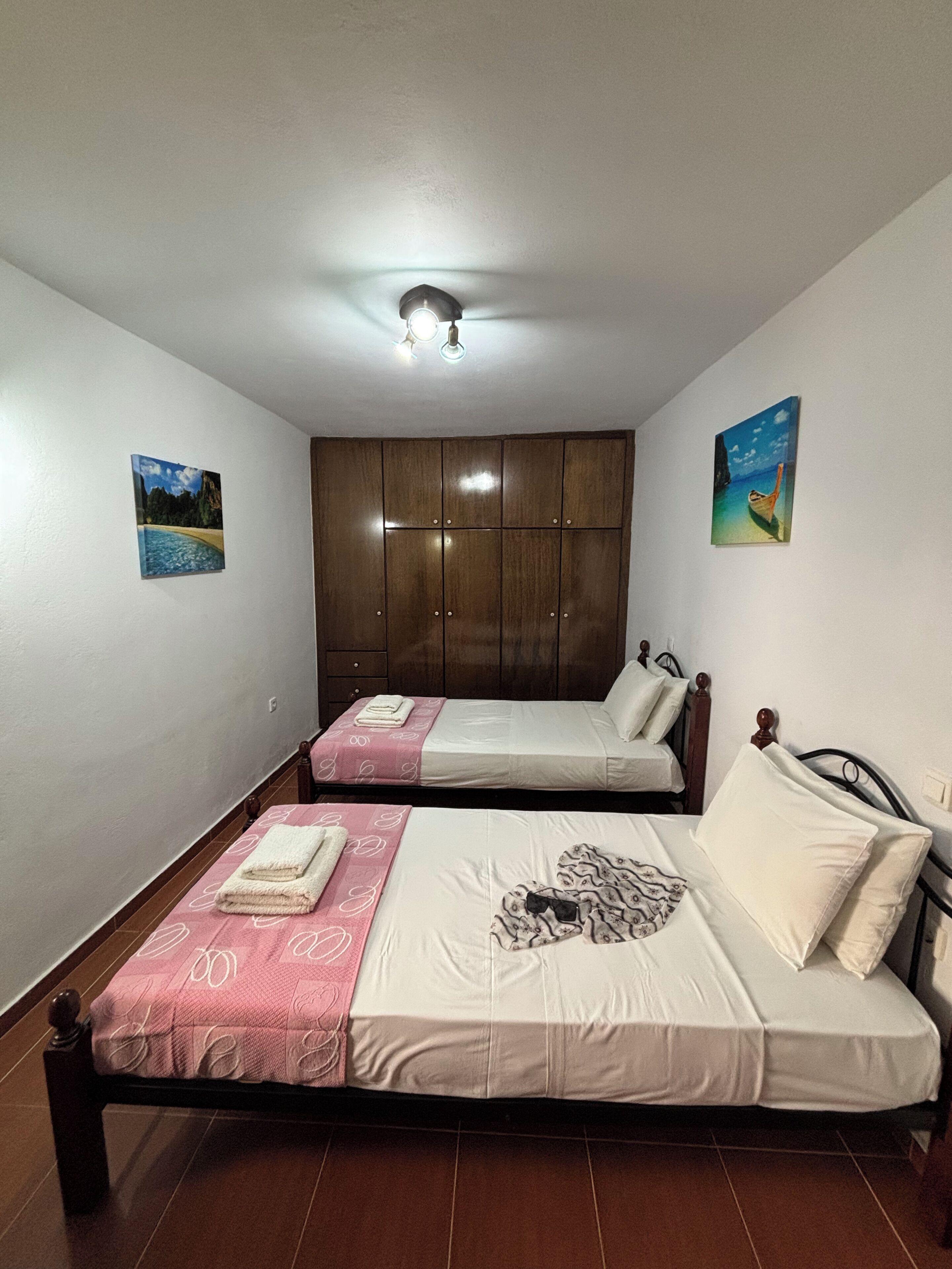 A bedroom with two beds, a wardrobe, and two paintings on the walls.
