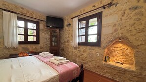 A bedroom with a stone wall, a fireplace, a bed with a pink blanket, and two windows with curtains. - Electra Stone Suite  (Roumeli)