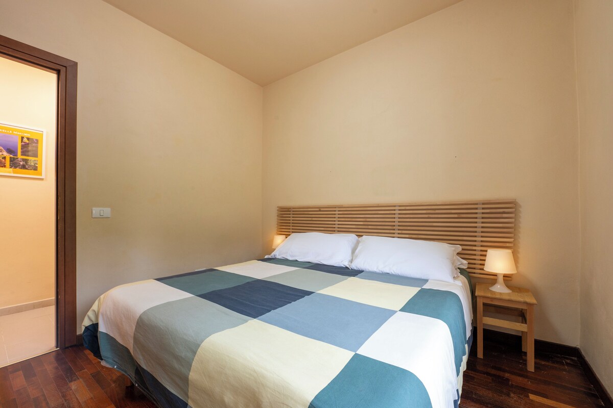 A bedroom with a bed, wooden headboard, bedside table with a lamp, and a framed picture on the wall.