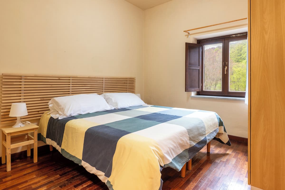 A bedroom with a wooden bed, a bedside table with a lamp, and a window with a view of trees.