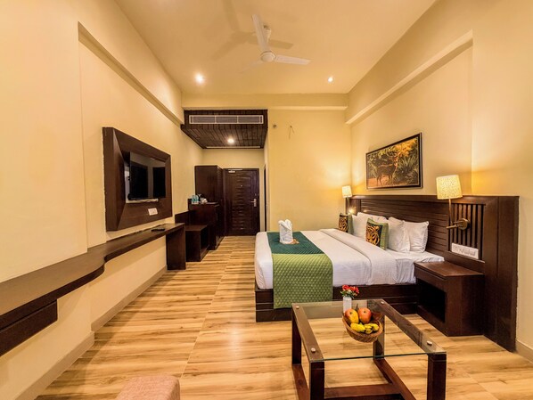 A hotel room with a bed, a TV, a desk, a chair, a glass coffee table with fruit, and a ceiling fan.