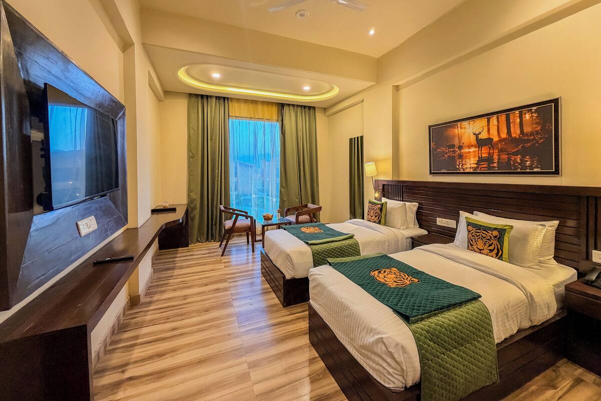 A hotel room with two beds, a TV, a ceiling fan, a painting, and a window with curtains.