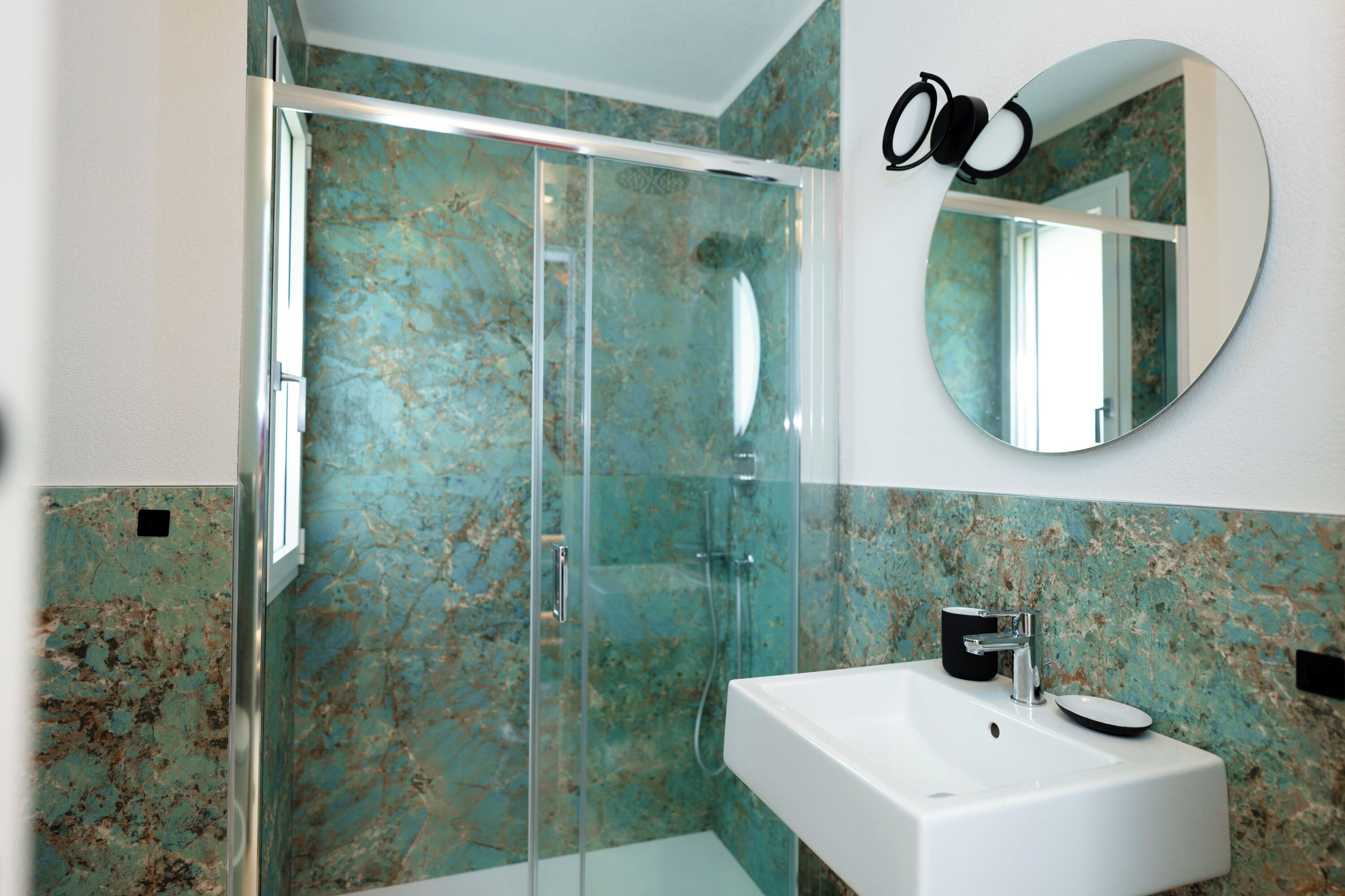 A bathroom with a glass-enclosed shower, a white sink, and a round mirror.