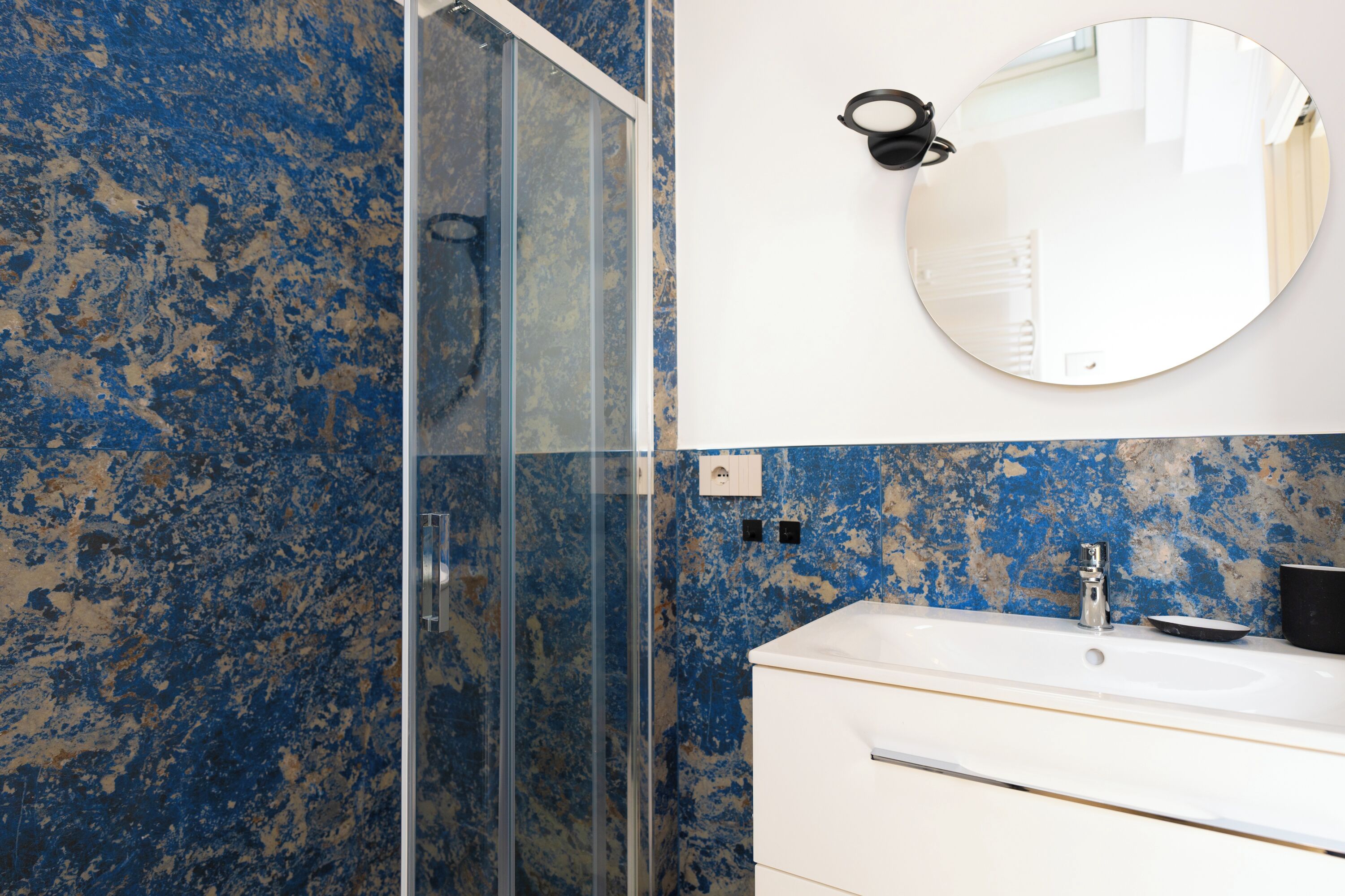 A bathroom with a glass shower enclosure, a white sink, and a round mirror on the wall.