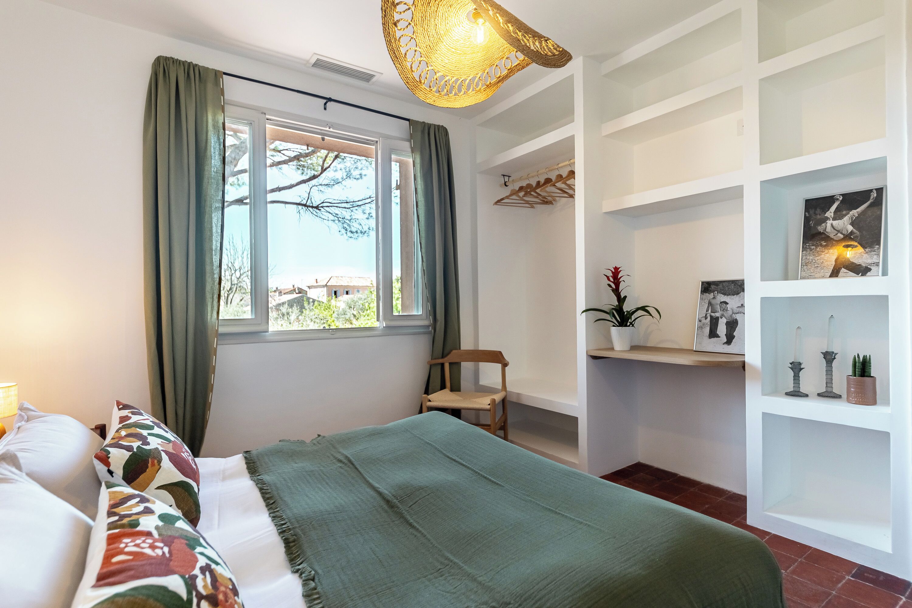 A bedroom with a bed, a chair, a window with curtains, a shelf with decorative items, and a hanging light fixture.