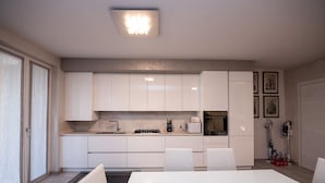 Een moderne keuken met witte kasten, een eettafel en stoelen. Er is een magnetron, een waterkoker en een spoelbak.